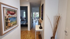 A hallway with a framed vintage poster, a small table with a vase, and a door leading to a living area with a sofa and a dining table. - One Level at Broadbeach - Inverloch (Inverloch)