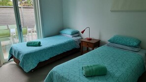 Une chambre avec deux lits, chacun recouvert d’une courtepointe bleue à motifs géométriques et agrémenté d’oreillers assortis. Une petite table de chevet en bois avec une lampe rouge est placée entre les deux lits. Une serviette verte est posée sur chaque lit. La chambre est dotée d’une porte-fenêtre coulissante qui donne sur un espace extérieur avec une terrasse en bois.