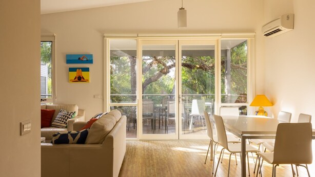 A modern living room with a sofa, a dining table with chairs, and a large sliding glass door leading to an outdoor area. - One Level at Broadbeach - Inverloch (Inverloch)
