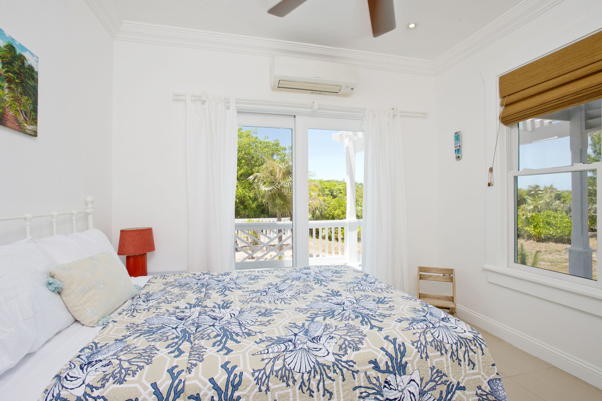 A bedroom with a large bed, a ceiling fan, an air conditioning unit, a window with a view of greenery, and a door leading to an outdoor balcony.