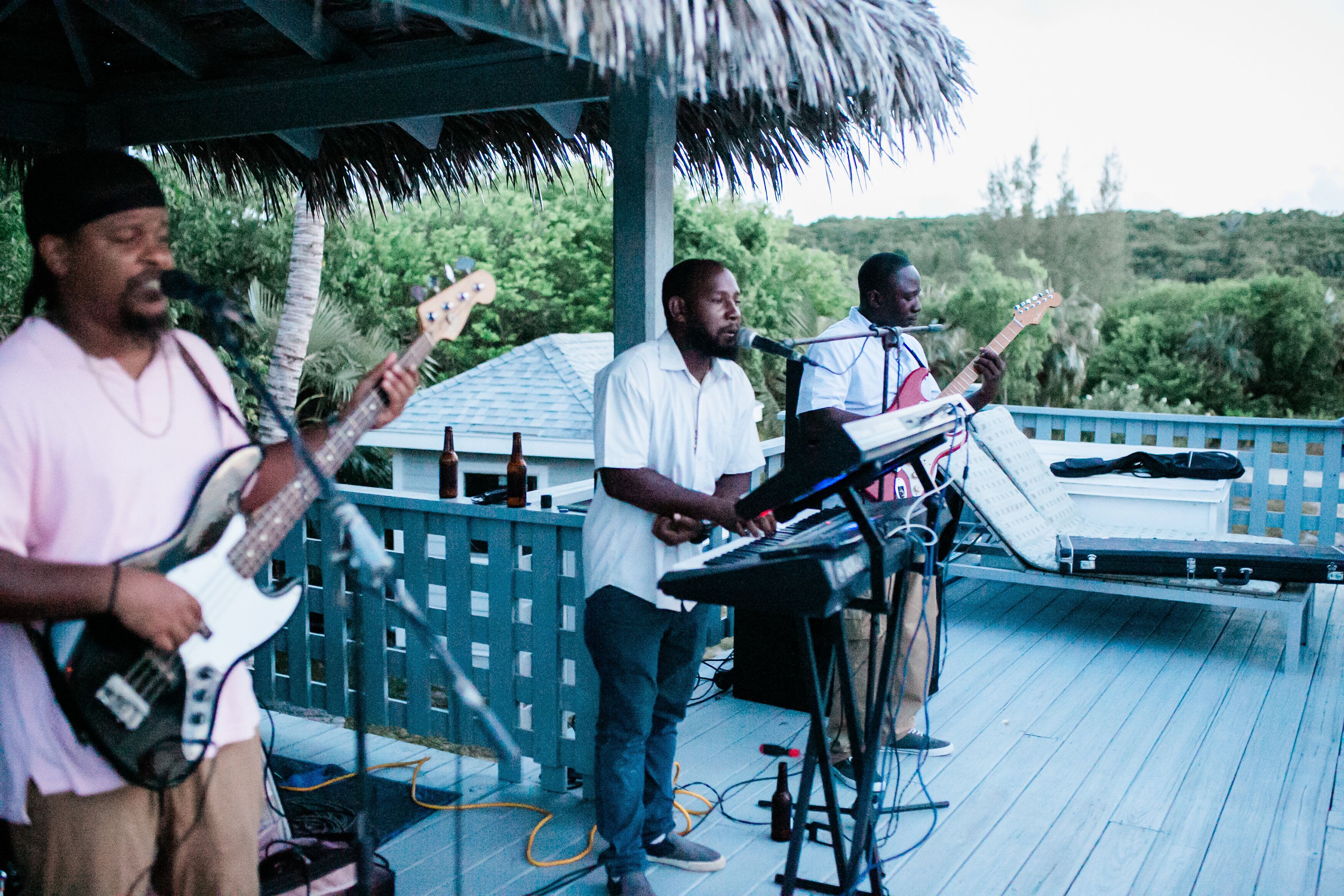 A band performing on an outdoor stage with a thatched roof, surrounded by lush greenery.