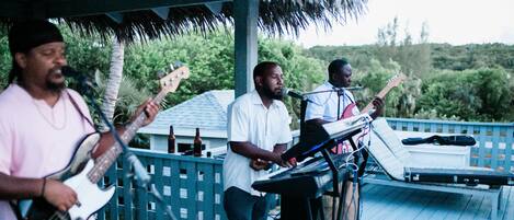 A band performing on an outdoor stage with a thatched roof, surrounded by lush greenery.