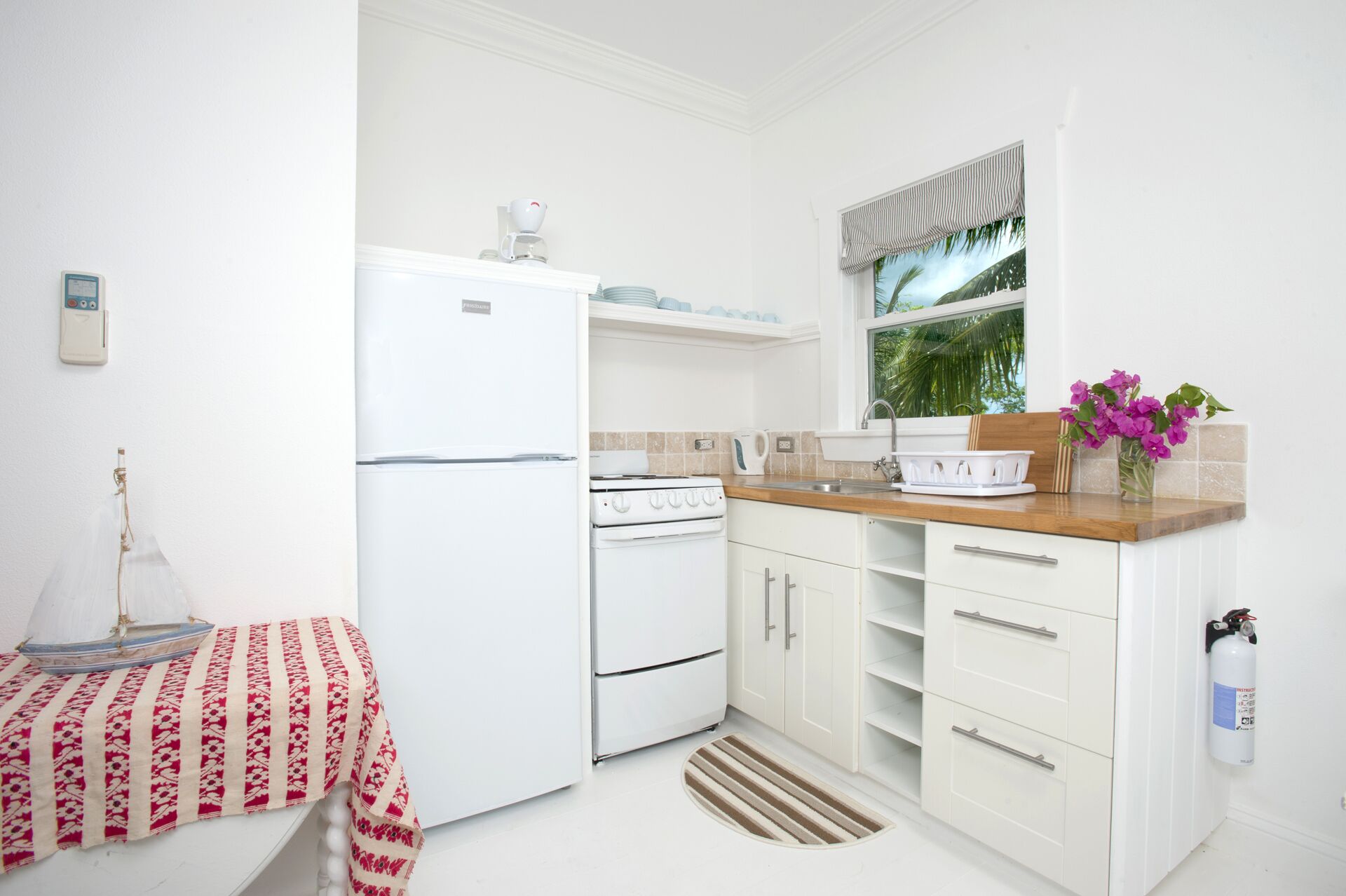 A compact kitchen with a refrigerator, stove, and sink. There is a window with a view of palm trees, a small table with a patterned tablecloth, and a vase with purple flowers.