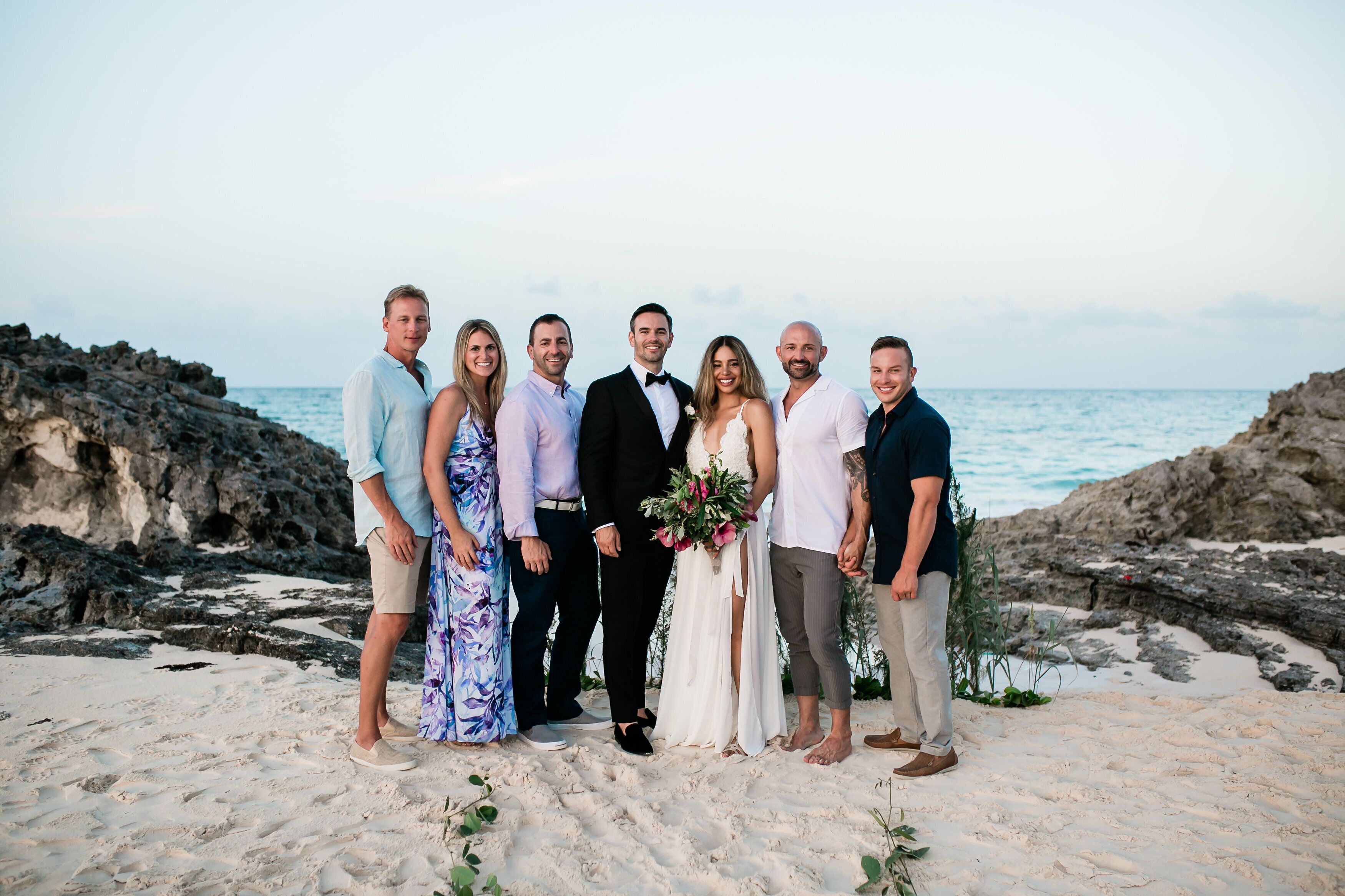 A group of people standing on a sandy beach with rocky formations and the ocean in the background.