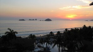 A sunset view over a beach with palm trees and a small boat in the water. - Amara Ixtapa, Luxury Oceanfront Condo, 6th Floor (Ixtapa)