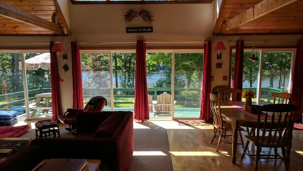 A spacious living area with a sofa, dining table, and chairs, large windows with red curtains, and a view of a lake and greenery. - Lakefront home with something to enjoy for every season! (Lee)