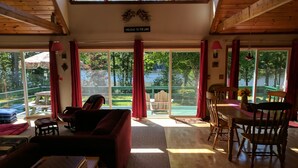 A spacious living area with a sofa, dining table, and chairs, large windows with red curtains, and a view of a lake and greenery. - Lakefront home with something to enjoy for every season! (Lee)