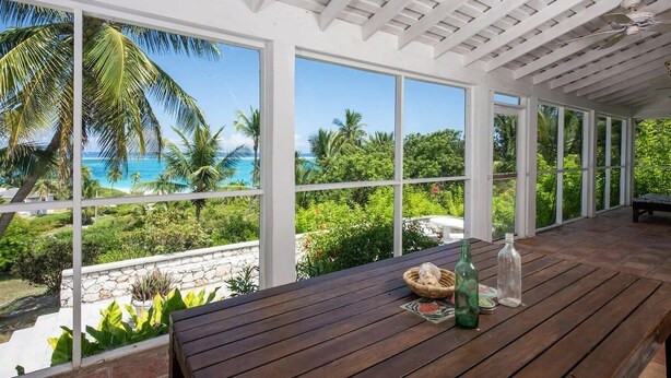 Un espace extérieur abrité, doté d’une table en bois, de grandes fenêtres et offrant une vue sur une plage et des palmiers.