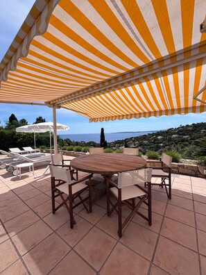 Eine Terrasse mit einem runden Holztisch und Stühlen, einem Streifenmarkisenstoff und Blick auf das Meer und die Hügel.