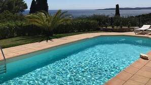 Ein klares, blaues Schwimmbecken mit einer gefliesten Kante, umgeben von viel Grün und Blick aufs Meer.
