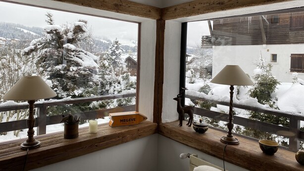 A corner room with wooden beams, a window with a view of snow-covered trees and a mountain, and a small table with a lamp and a decorative item.