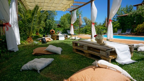 Un espace extérieur aménagé avec des coussins blancs et beiges, une table en bois et une piscine en arrière-plan.