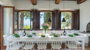 Comedor con una mesa larga dispuesta para seis comensales, rodeado de ventanas grandes con contraventanas de madera, que ofrece vistas a zonas verdes y colinas.