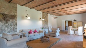 Amplia sala de estar con pared de piedra, techo de madera y comedor con mobiliario blanco.