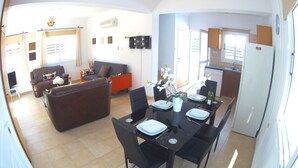 Un salon avec un canapé, une table à manger dressée pour deux personnes et un coin cuisine équipé d’un réfrigérateur.