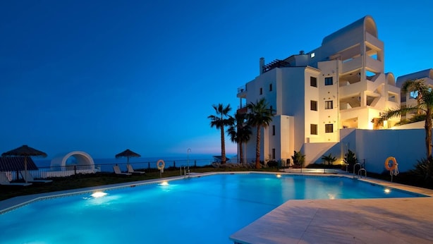 Une piscine avec vue sur la mer, un bâtiment moderne doté de balcons et bordé de palmiers.