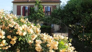 A building with a tiled roof and arched windows, surrounded by lush greenery and flowering plants. - Lovely Barn with Private Poolnear San Gimignano, between Florence and Siena. (Castelfiorentino)