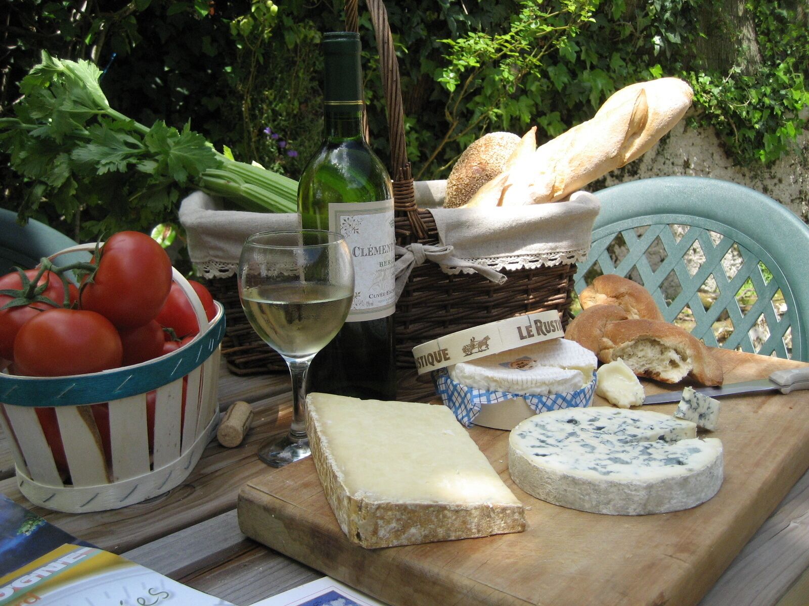 A wooden table with a bottle of CLEMENT B. BERLIN wine, a glass of white wine, a basket of tomatoes, a loaf of bread, a block of cheese, and a blue cheese wheel.