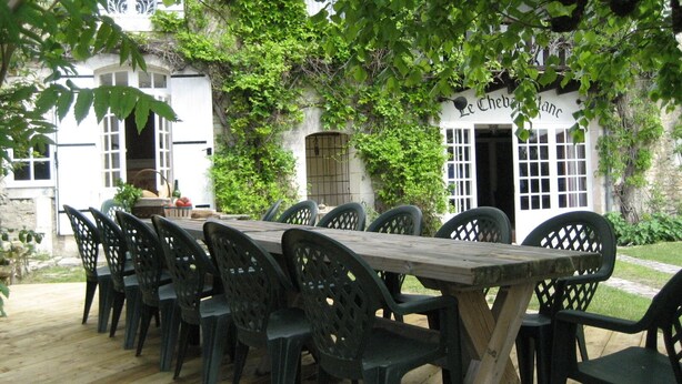 Une table en bois installée à l’extérieur, avec des chaises, entourée de verdure et d’un bâtiment sur lequel on peut lire l’inscription « Le Cheval ».