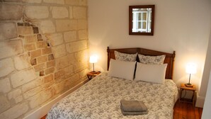 Une chambre avec un mur en briques, un lit en bois avec une literie à motifs floraux, deux lampes de chevet et une petite fenêtre au-dessus du lit.