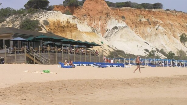 Una playa con un acantilado rocoso al fondo, un área de descanso con sombrillas y un sendero arenoso.