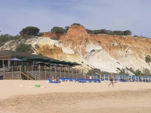 Ein Strand mit einer felsigen Klippe im Hintergrund, einem überdachten Sitzbereich mit Sonnenschirmen und einem Sandweg.