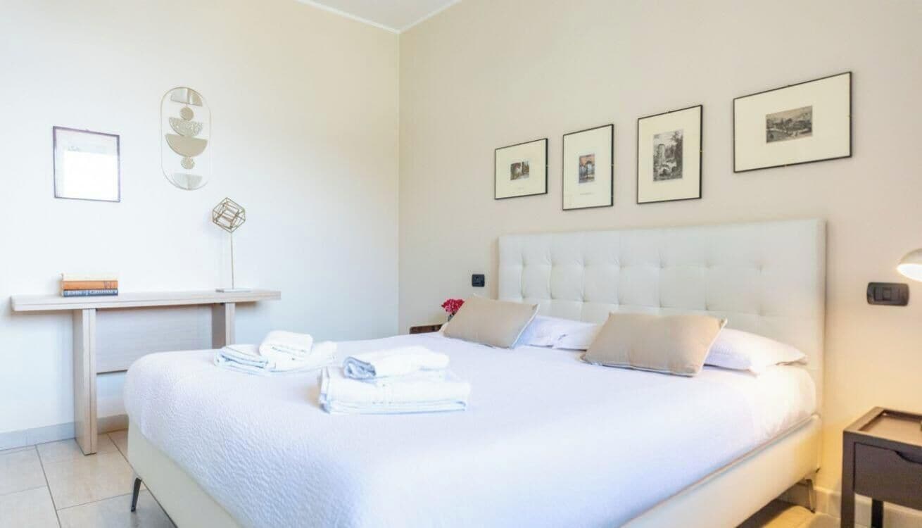 A neatly made bed with pillows and folded towels, a bedside table with books, and a wall-mounted picture frame.