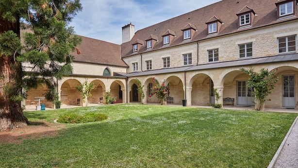 Une cour intérieure avec un bâtiment en pierre, dotée d’arcades et d’une pelouse verdoyante.