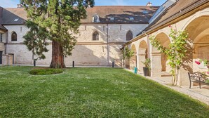 Une cour avec un grand arbre, des bâtiments en pierre et un banc.