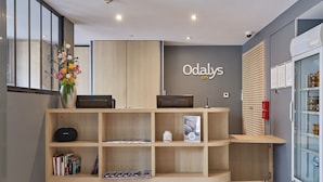Une salle d’attente avec un bureau en bois, un vase rempli de fleurs, un réfrigérateur et une enseigne fixée au mur portant l’inscription « Odaly’s ».