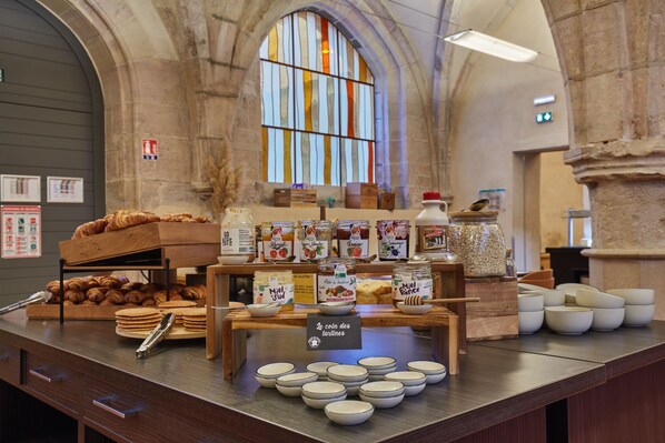 Mostrador de panadería con pasteles, frascos de conservas y tazones de crema en un edificio histórico de paredes de piedra y ventanal de vidrio emplomado.