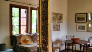 Une pièce dotée d’un grand miroir décoratif qui reflète un lit avec un oreiller, une fenêtre avec des stores, une table à manger avec des chaises et des tableaux encadrés accrochés au mur.