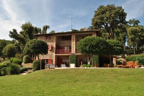 A two-story stone house with a balcony, surrounded by lush greenery and trees.
