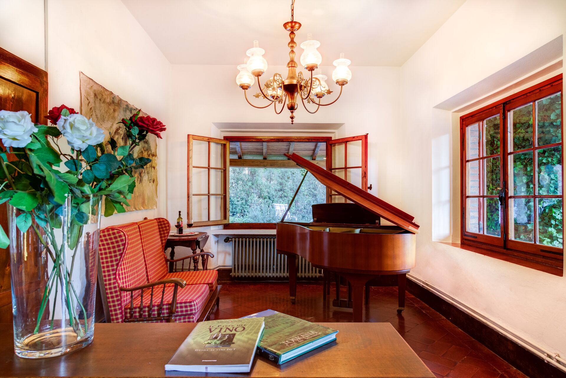 A room with a grand piano, a red and white checkered sofa, a table with books, a vase with flowers, and a large window with a view of greenery.