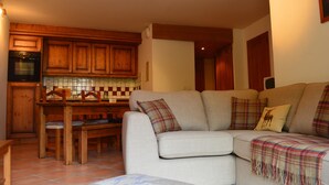 Ein gemütliches Wohnzimmer mit einem beigefarbenen Sofa, einem Holzesstisch und einer Küchenzeile mit Holzschränken und einer gefliesten Rückwand.
