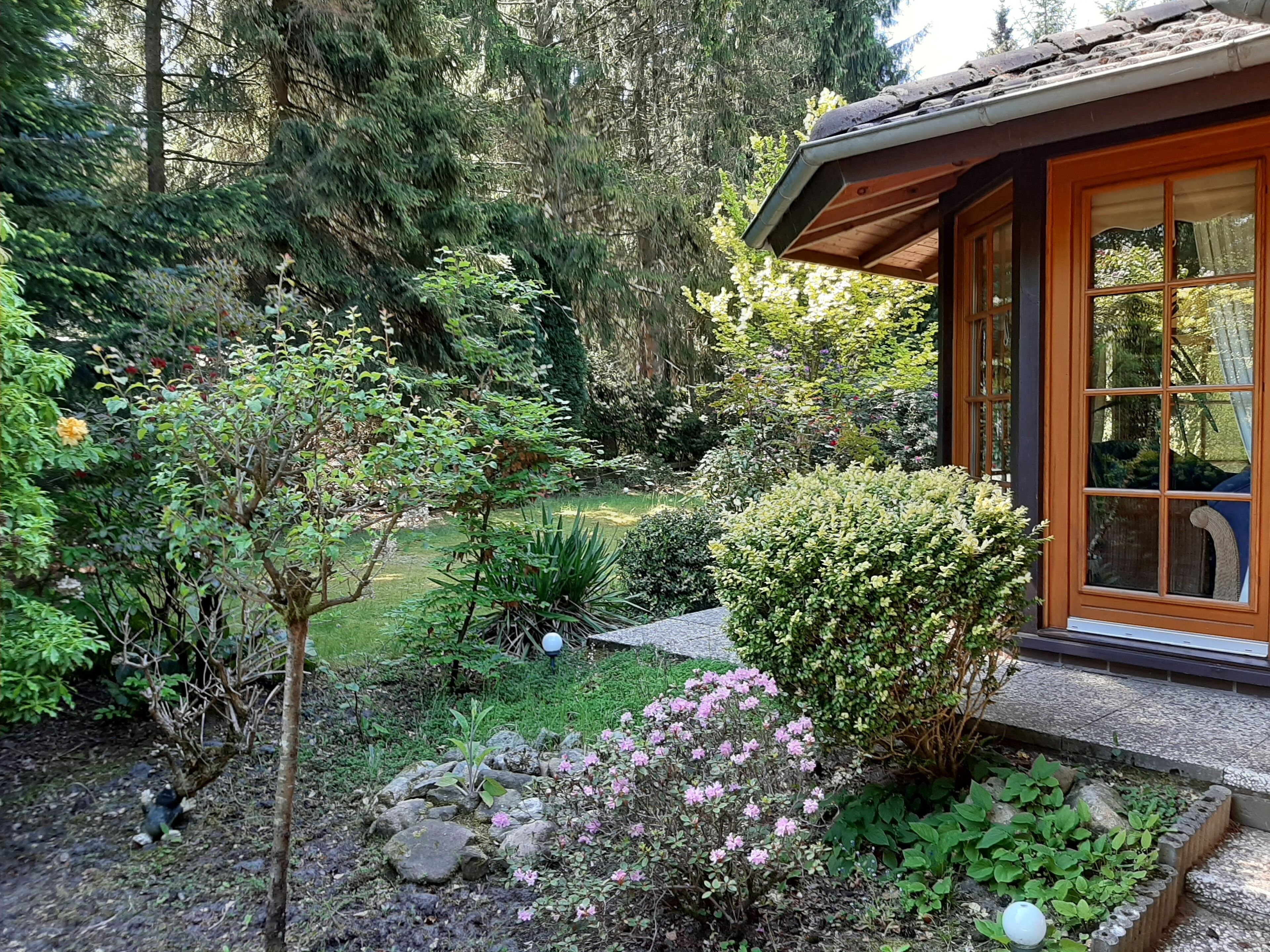 Eine Holzhütte mit steinernen Fundamenten, umgeben von einem üppigen Garten mit verschiedenen Pflanzen und Blumen.