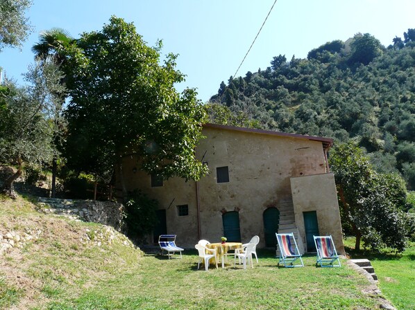 A rustic building with a sloped roof, surrounded by lush greenery and hills. There are outdoor seating arrangements with tables and chairs. - Nice small house with garden and breath-taking view over the Versilia Coast (Monteggiori)