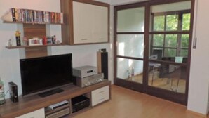 Ein Wohnzimmer mit Flachbildfernseher, Bücherregal und einer Schiebetür zu einem Balkon.