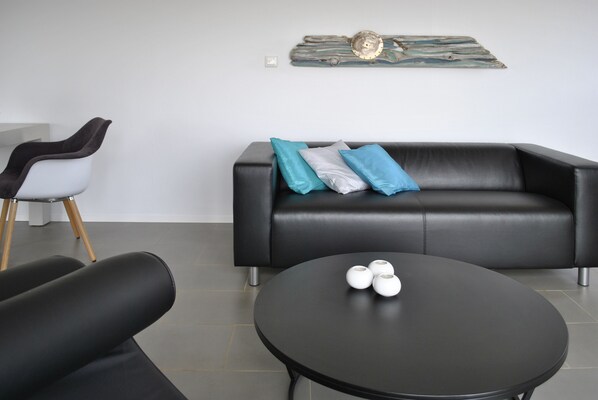 Ein modernes Wohnzimmer mit einem schwarzen Ledersofa, einem runden schwarzen Couchtisch und einem weißen Sessel mit braunem Kissen.