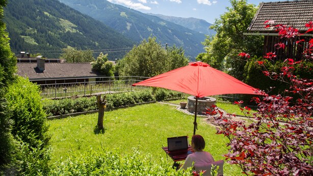 Une personne utilise un ordinateur portable sous un parasol rouge, dans un jardin luxuriant avec vue sur la montagne.