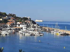 Una marina costiera con numerose imbarcazioni ormeggiate, una grande nave da crociera in lontananza e edifici lungo la riva.