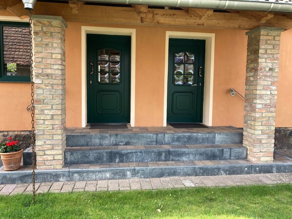 A house with two green doors, a brick exterior, and a small porch with steps leading to the entrance.