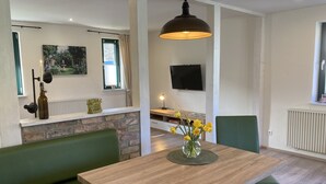 Een moderne woonkamer met een houten eettafel, groene bekleding, een accentmuur van steen, een tv aan de muur en een hangende hanglamp.
