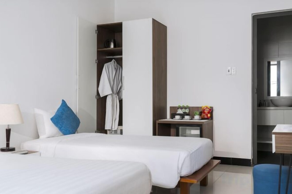 A hotel room with two beds, a wardrobe, a desk with a lamp, and a bathroom visible through an open door.