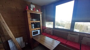 A cabin interior with a wooden table, a built-in shelf with kitchen appliances, a window with a view of a snowy landscape, and a bench with red cushions. - Furnished studio rental Winter/Summer Savoie ski area at the foot of the slopes (Villarembert)