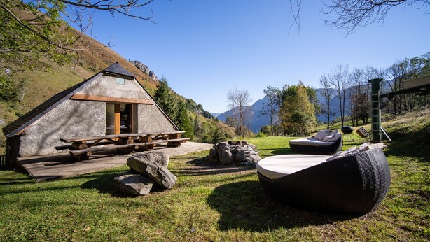 Eine rustikale Holzhütte mit einer Holzdiele und einem Kamin aus Stein, umgeben von einer grünen Wiese und Blick auf die Berge.