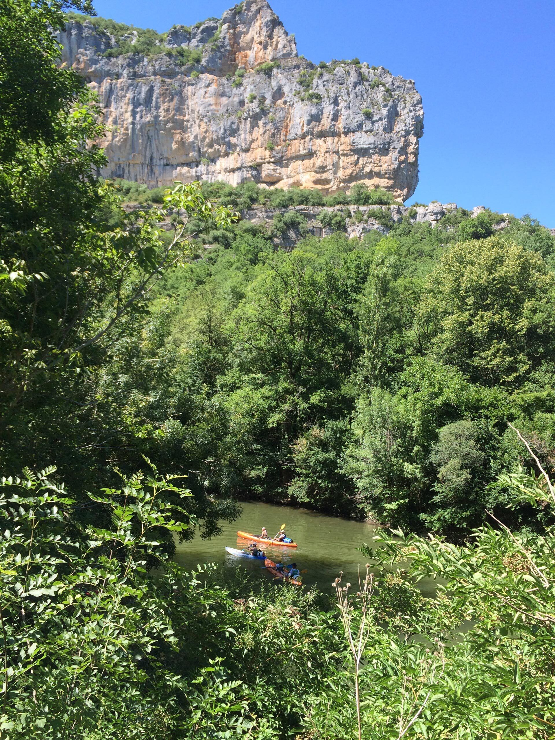 A river with kayaks, surrounded by dense greenery and a rocky cliff in the background.