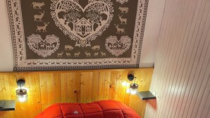 Ett rum med en sänggavel i trä, ett rött sängöverkast och en vävtapet med hjärt- och djurmotiv.