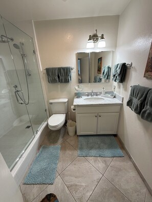 Et badeværelse med et glasafskærmet brusebad, toilet, vask med spejl og to håndklæder hængende på væggen.