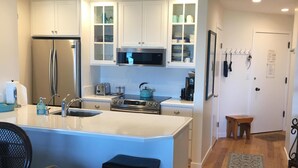 A modern kitchen with white cabinets, a stainless steel refrigerator, a microwave, and a stove. There is a small island with a sink and stools. - Magical Place By The Sea. (Watsonville)